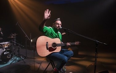 Paweł Domagała na scenie z gitarą i witający publiczność podnosząc otwartą dłoń ku g&oacute;rze